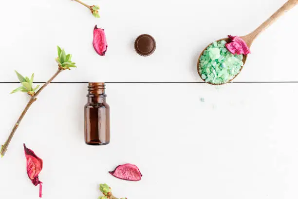 Dark bottle with essential oil, dry flowers of a rose and green sea salt, on a white wooden table. Top view. Copy space