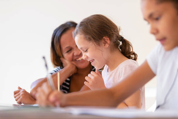 Young Aboriginal girls studying with their mother helping. They are working at a table at home. One girls listening to music or educational work as she works. They are sisters doing schoolwork together with assistance from mum.