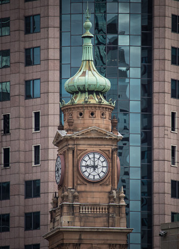 An old clock tower appears in front of some modern tall office buildings, which overshadow the vintage and historic building.