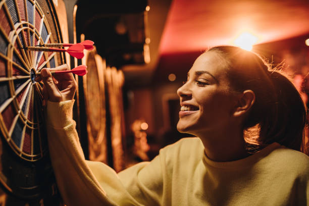 Young happy woman taking darts out of dart board. Young smiling woman removing darts out of dart board in entertainment club. woman darts stock pictures, royalty-free photos & images