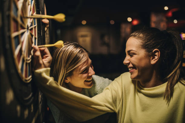 Happy women talking while removing darts from dartboard. Two female friends having fun while taking darts out of dartboard in a bar and talking. dart stock pictures, royalty-free photos & images