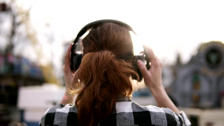 Backside footage of a girl with a brown haired tail standing a plaid shirt oover against the shining sun. Putting on the headphones