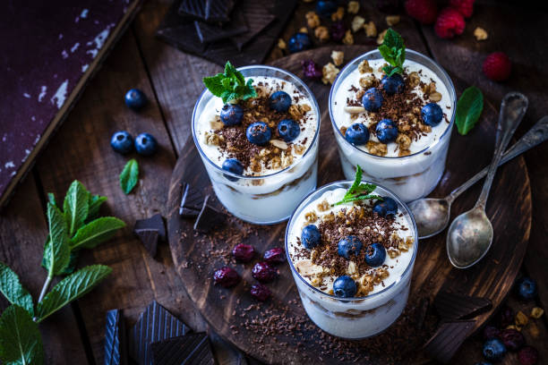 Yogurt with granola, berry fruits and chocolate High angle view of three glasses of homemade yogurt with granola, berry fruits and chocolate shot on rustic wooden table. The glasses are on a round wooden tray and two spoons are also on the tray. Low key DSRL studio photo taken with Canon EOS 5D Mk II and Canon EF 100mm f/2.8L Macro IS USM jar of milk stock pictures, royalty-free photos & images