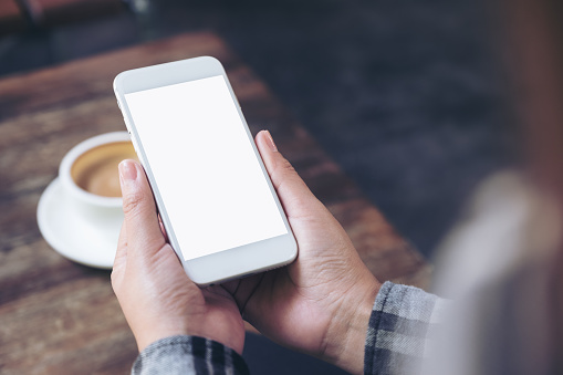 Mockup image of a woman's hands holding white mobile phone with blank desktop screen with coffee cup on wooden table in cafe