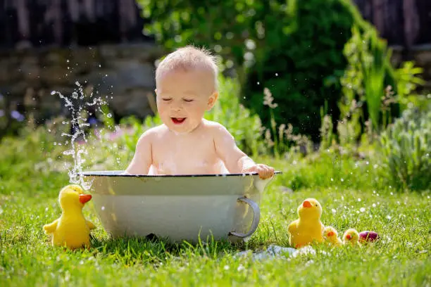 Photo of Cute little toddler boy in a basin, taking a bath in garden with bubbles and duck toys