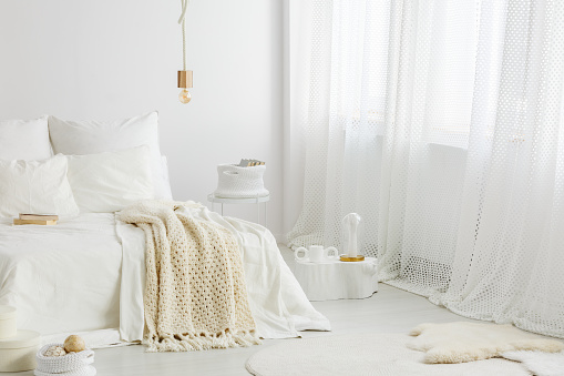 Cozy, knit blanket on white bed in bright bedroom interior with a big window