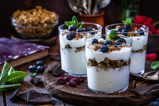 Three glasses of homemade yogurt with granola, berry fruits and chocolate shot on rustic wooden table. The glasses are on a round wooden tray and two spoons are also on the tray. Low key DSRL studio photo taken with Canon EOS 5D Mk II and Canon EF 100mm f/2.8L Macro IS USM
