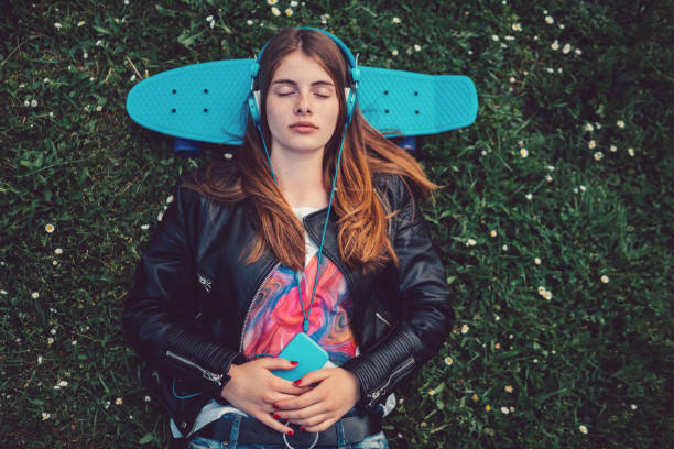 Relaxed teenage girl in the grass listening to music Young woman lying down in the grass with skateboard behind head mindfulness kids stock pictures, royalty-free photos & images