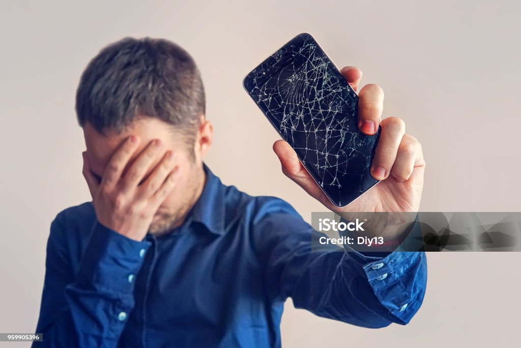 The guy is holding a black smartphone with a broken display. The guy is holding a black smartphone with a broken display. Broken screen of modern frameless phone. guy crying with his eyes closed and hands in his face. Broken Stock Photo