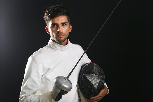 Young man in fencing equipment - uniform, face mask and foil sword on black background. About 20 years old, Caucasian or Latin man.