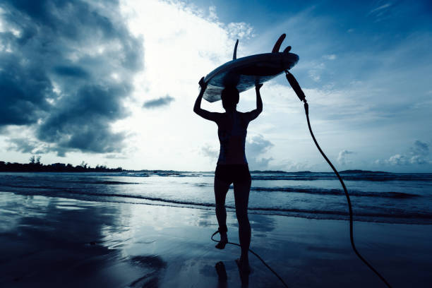 Silhouette of a man holding a surfboard at dawn. He is looking at the ocean waves with beautiful sunrise colours in the sky.