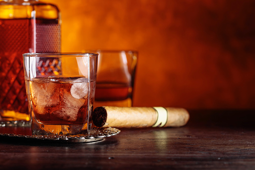 Glass of scotch whiskey with natural ice and cigar on old wooden table.