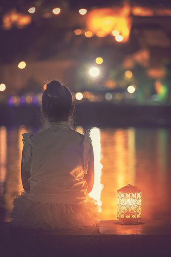 Beautiful little girl holding a lantern and enjoy on night view of city while sitting on promenade near waterfront