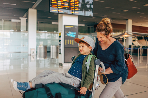 Photo of a cheerful little boy and his mother, who travel together, pushing a luggage cart at the airport