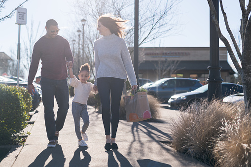 A pre-school age tugs her parents along outside the grocery store. They are outside on the sidewalk and she is holding their hands. Mom is carrying a paper bag of groceries. It's a sunny day.