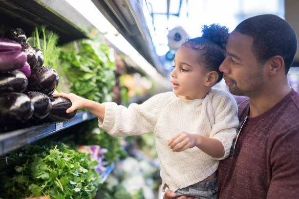 Dad takes his young daughter grocery shopping A pre-school age girl helps her dad pick out veggies in the produce section at the grocery store. He is holding her next to the produce and she is picking out eggplant. healthy groceries stock pictures, royalty-free photos & images
