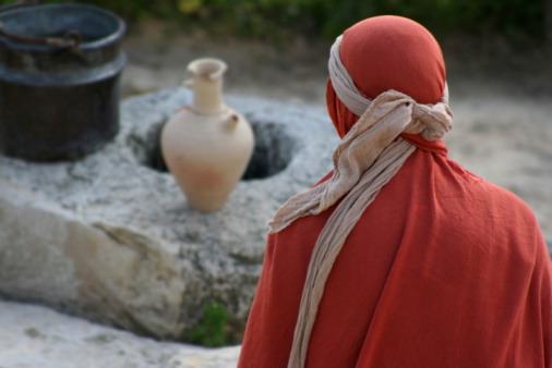 A woman wrapped in cloth sits in front of a 1st century well.