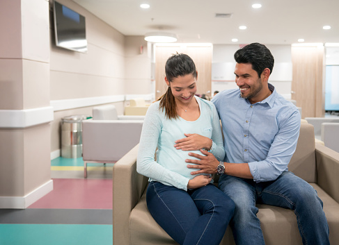 Beautiful young couple sitting at the waiting room of the hospital while husband touches her belly waiting for consult with obstetrician both smiling