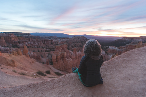 An ethnic senior woman sits on a cliff in the remote Utah desert and watches the sun set. She is alone. There is a whole range of mountains spread out before her. The shot is from behind.