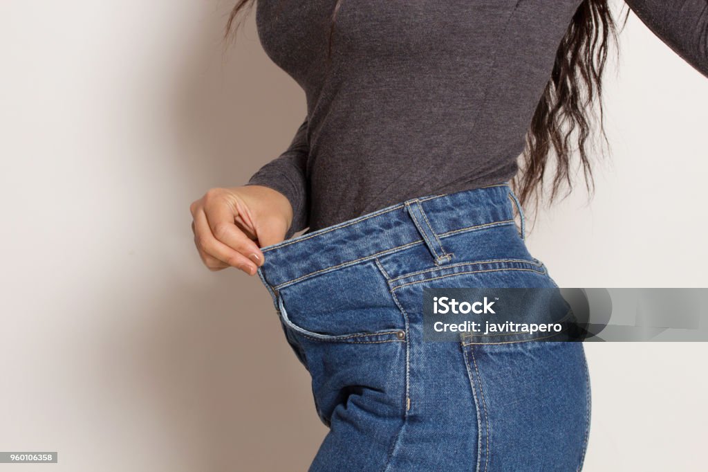 Latin woman in baggy pants, indoors. Latin woman in baggy pants, indoors, over a white wall. Weight loss concept.Latin woman in baggy pants, indoors, over a white wall. Weight loss concept. Liposuction Stock Photo