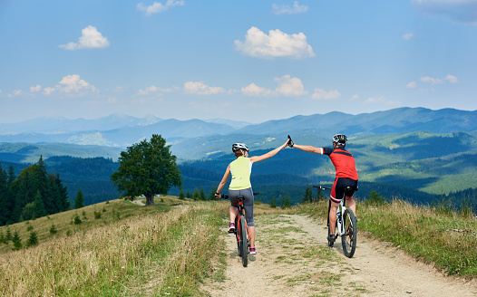 Back view of active couple cyclists in professional sportswear and helmets cycling down cross country bikes on the mountain road on summer day. Sporty man and woman giving each other a high five