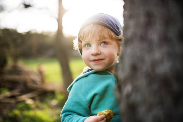 un garçon heureux bambin se cachant derrière l’arbre à l’extérieur dans la nature du printemps. - tree grass family human relationship family photos et images de collection