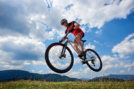 Young athletic sportsman biker in professional sportswear flying in air on his bike on bright blue sky with white clouds and distant hills background Active lifestyle and extreme sport concept.