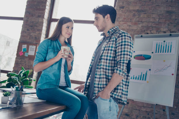 pretty girl sitting on desktop holding cup of tea, near  standing stylish man, holding hand in pocket, flirting, couple looking to each other in work station - staring imagens e fotografias de stock