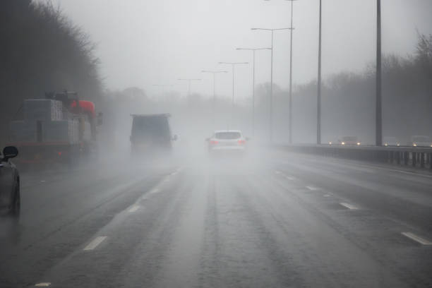 Several cars on the road are about to enter a dense fog. A danger sign warns of an approaching right turn. There are snow drifts