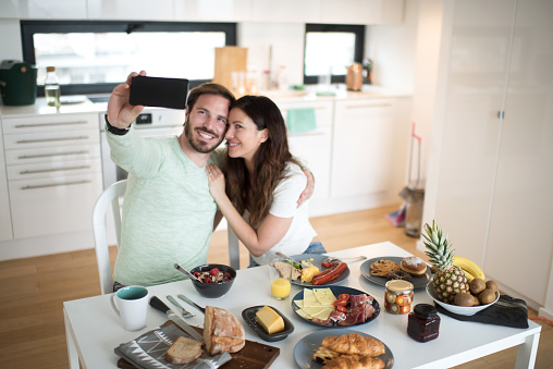 Heterosexual couple in their new apartment having a meal together and posing for a selfie image.