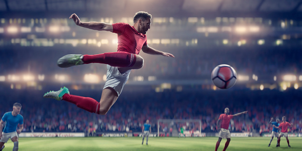 A professional male soccer in mid air with arms out and legs back, about to kick the ball in a volley. The footballer is wearing generic red and white soccer kit and is playing in a soccer match in a generic outdoor stadium. With selective focus and background bokeh.