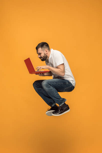 若い男がジャンプしながらノート パソコンを使用してオレンジ色の背景上のイメージ - beautiful business lifestyles studio shot ストックフォトと画像