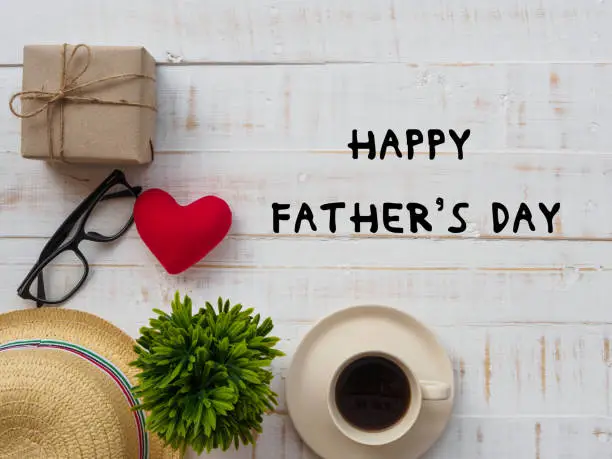 Happy Father's Day inscription with redheart, gift box, hat, glasses, plant and a cup of coffee  on white wooden background.father's day concept.