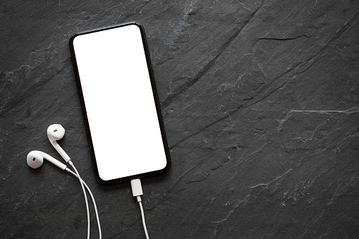 Mobile phone with empty screen and earphones on black stone surface; mock-up for your design