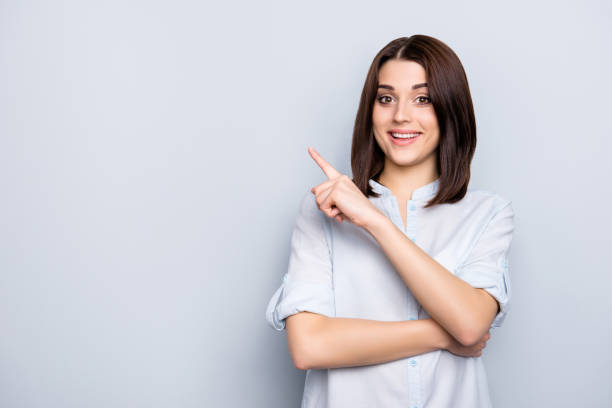 portrait with copy space of charming, pretty, nice, stylish, brunette, trendy woman in shirt with modern hairstyle, pointing  forefinger on empty place, looking at camera, isolated on grey background - blank sale young women one young woman only imagens e fotografias de stock
