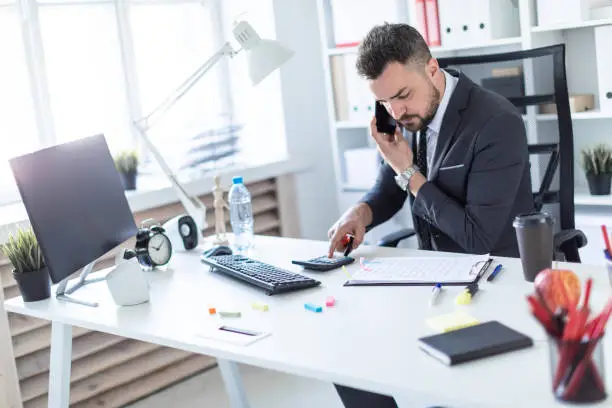 Photo of A man is sitting at the desk at the office, talking on the phone, holding a marker in his hand and counting on a calculator.