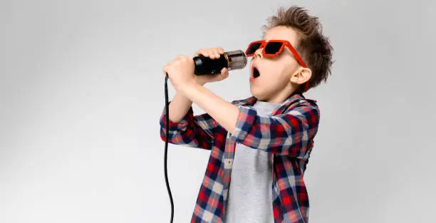 Charming happy child on gray background. The boy's hair is up. The boy has a hairstyle. The boy in round sunglasses. A handsome boy with a red hair color. The boy sings into the microphone.