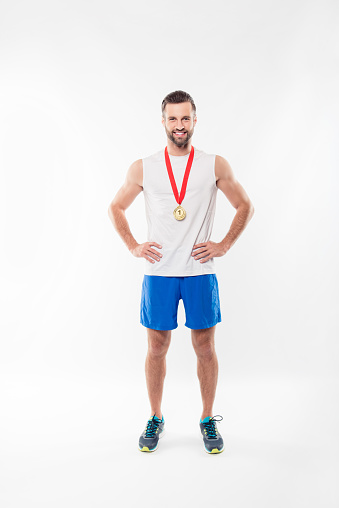 Full length, fullbody portrait of attractive, powerful, lucky, sporty, sexy, strong, athletic man having gold medal with red ribbon on his neck, hold arms on waist, isolated on white background