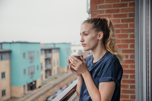 Side view of a young female adult standing out on a balcony and warming up with a cup of tea.