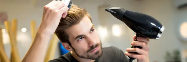 Male hairdresser combing and drying his own hair in hair salon in front of the mirror.