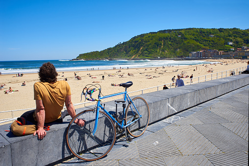 San Sebastian, Spain - May 11, 2018. A young men with their bike parked in front of  Zurriola beach and Monte Ulia in background at sunny day. San Sebastian, Guipuzcoa. Spain.