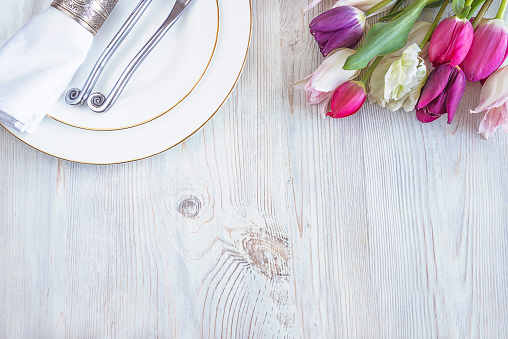 Festive table setting with original cutlery on white napkin and bouquet of tulips on white wooden background with copy space