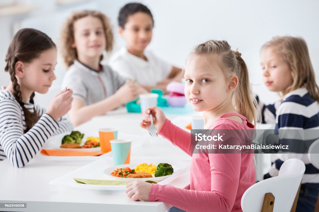 Girl eating vegetables with friends in the canteen during break at school Cafeteria Stock Photo