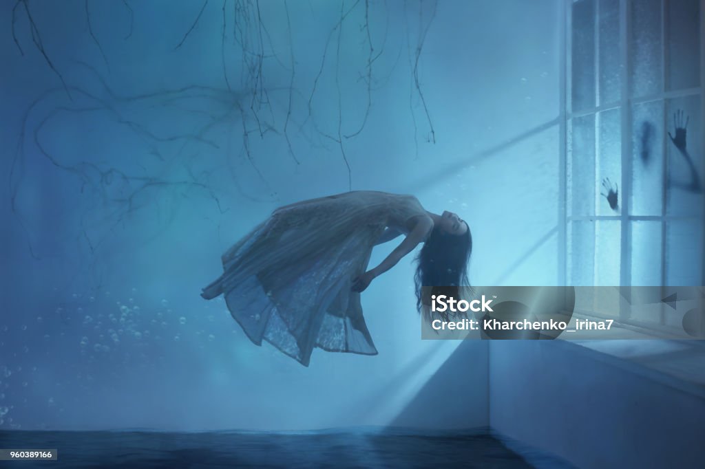A ghost girl with long hair in a vintage dress. Room under water. A photograph of levitation resembling a dream. A dark Gothic interior with branches and a huge window of flooded light. Art photo Ghost Stock Photo