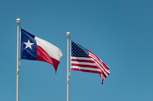 The state flag of Texas and American flag waving in the wind on flagpole. Blue sky background with copy space.