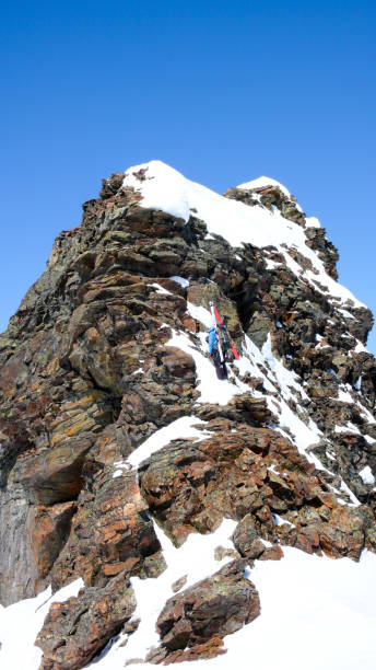 männliche backcountry skifahrer mit seinen ski an den rucksack geschnallt zu einem exponierten felsigen gipfel klettern - back country skiing extreme skiing skiing ski stock-fotos und bilder