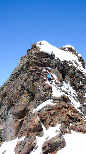 männliche backcountry skifahrer mit seinen ski an den rucksack geschnallt zu einem exponierten felsigen gipfel klettern - back country skiing extreme skiing skiing ski stock-fotos und bilder