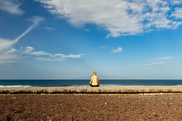 Woman meditating in yoga pose, ocean view, beach and wooden sidewalk. Motivation and inspirational summer sea landscape. Healthy lifestyle outdoors in nature concept.