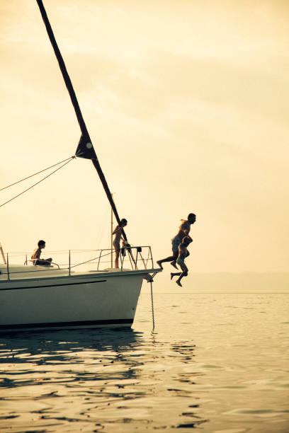 Parent having a fun with their children on vacation on sailboat. They jumping into the water from yacht bow. Sea is very nice and calm and sunset in background makes beautiful colors and reflections