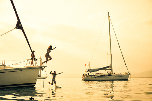 Parent having a fun with their children on vacation on sailboat. They jumping into the water from yacht bow. Sea is very nice and calm and sunset in background makes beautiful colors and reflections